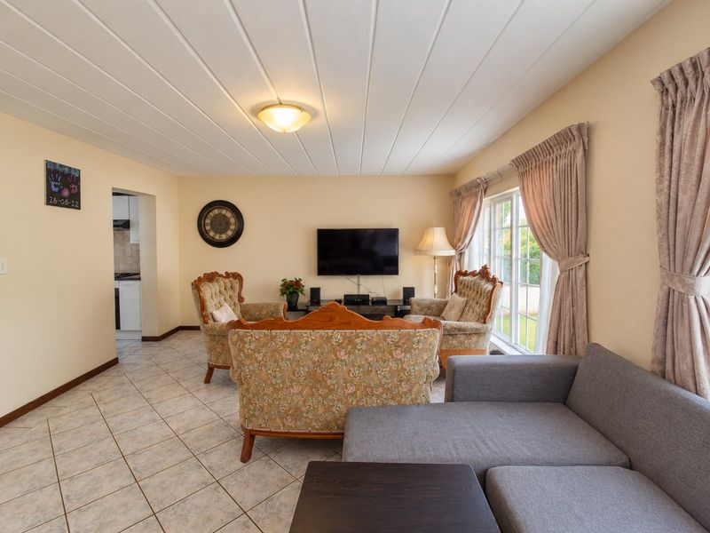 A spacious living room featuring tiled flooring, a panelled ceiling, and a large window allowing natural light, with an adjacent view into the kitchen area.