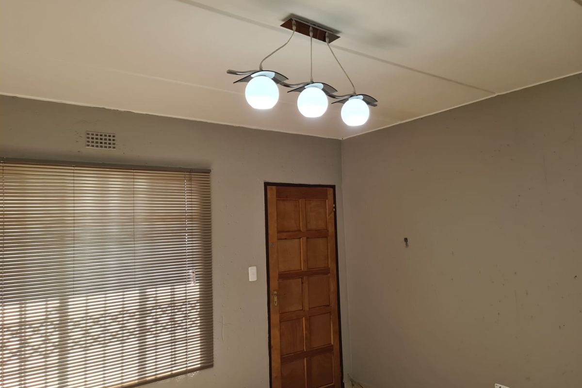 An empty living room featuring tiled flooring, a wooden door, venetian blinds, and a modern three-bulb ceiling light fixture.
