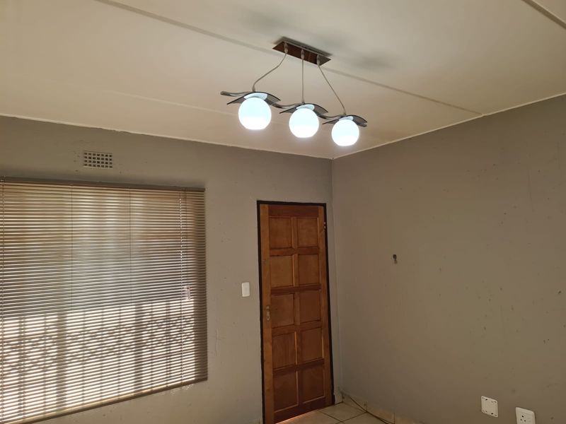 An empty living room featuring tiled flooring, a wooden door, venetian blinds, and a modern three-bulb ceiling light fixture.
