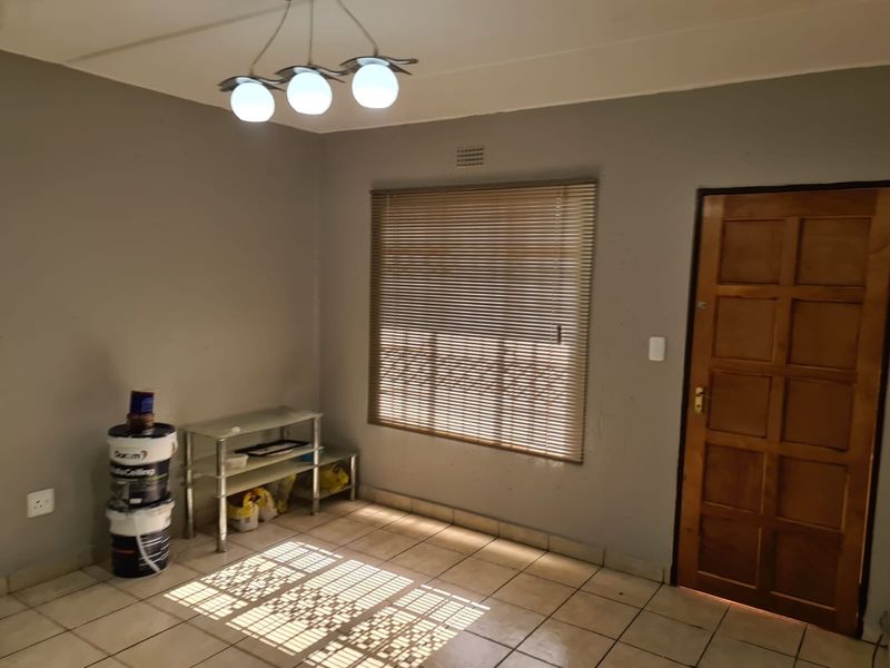 A sparsely furnished living area featuring tiled flooring, a wooden panel door, and natural light filtered through venetian blinds.