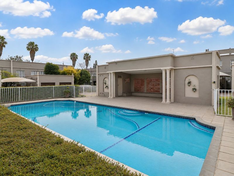 A well-maintained outdoor pool area featuring a clear swimming pool, paved deck, and a covered patio space with decorative architectural columns.