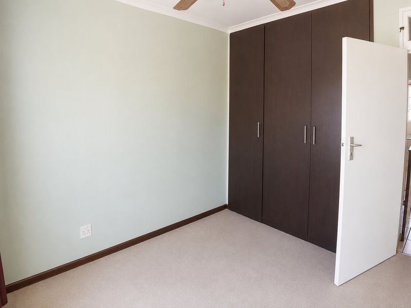 A clean, empty bedroom featuring neutral carpeting, built-in dark wood cupboards, and a ceiling fan, with a view into the adjacent hallway.