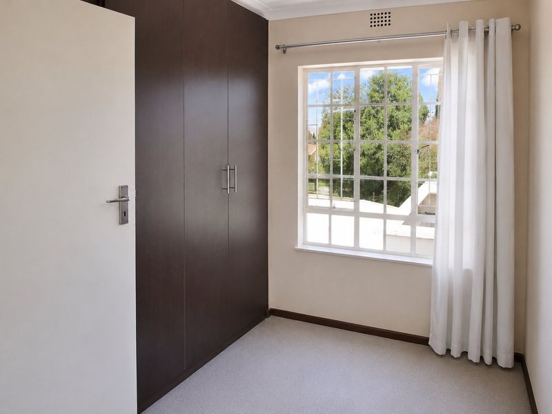 A bright, empty bedroom featuring dark wood built-in cupboards, carpeted flooring, and a window with security bars overlooking the exterior.