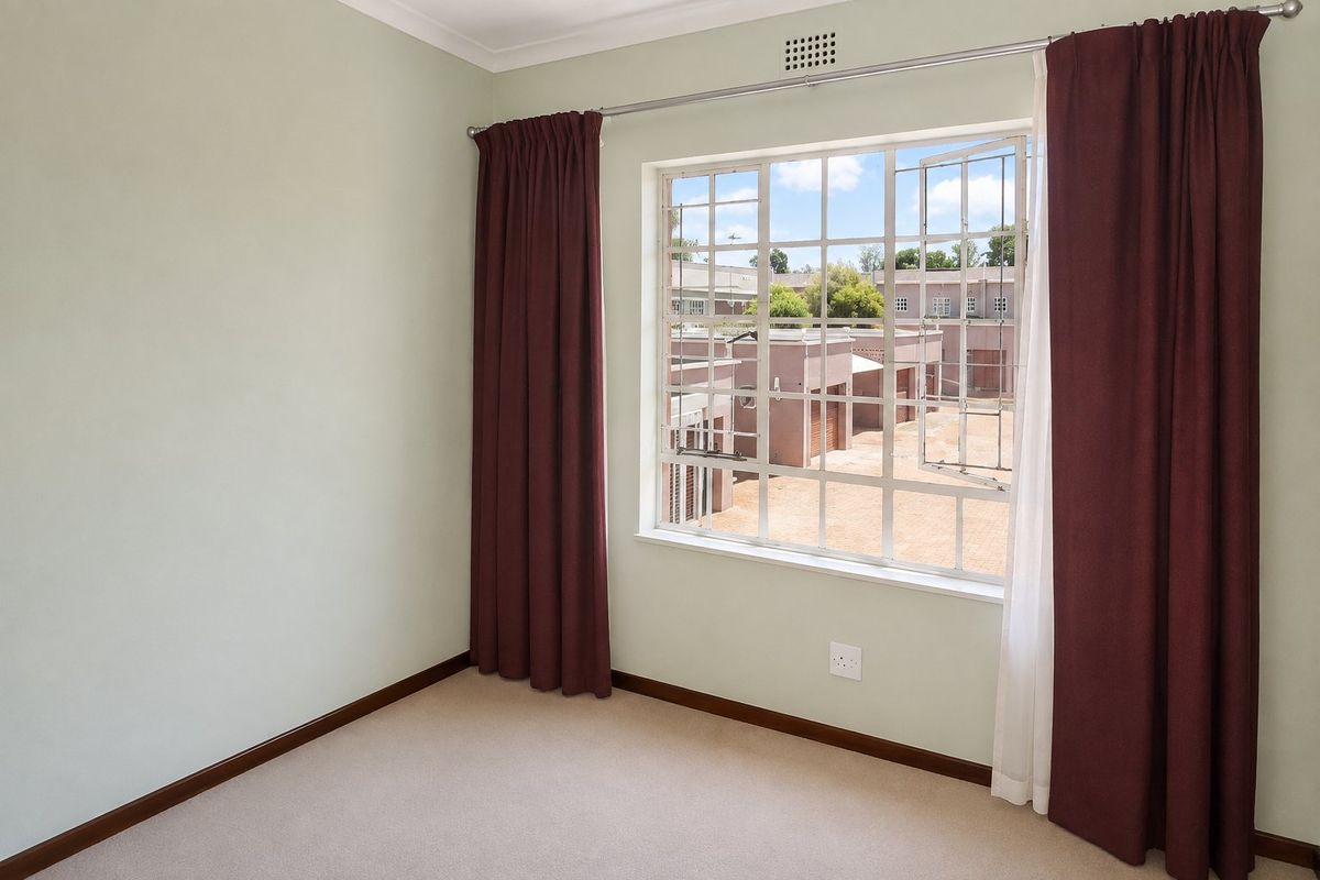A bright, empty bedroom featuring neutral carpeting, pale green walls, and a large steel-framed window overlooking the complex.