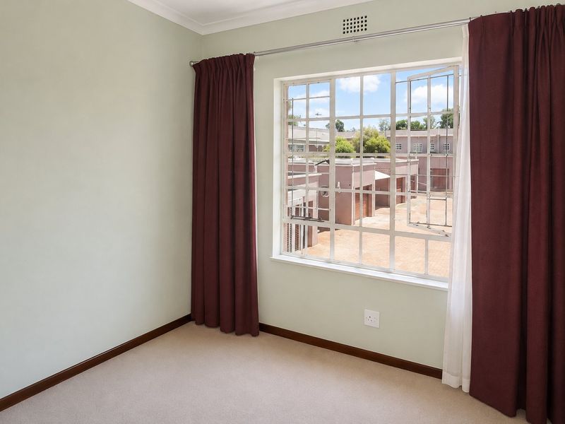 A bright, empty bedroom featuring neutral carpeting, pale green walls, and a large steel-framed window overlooking the complex.