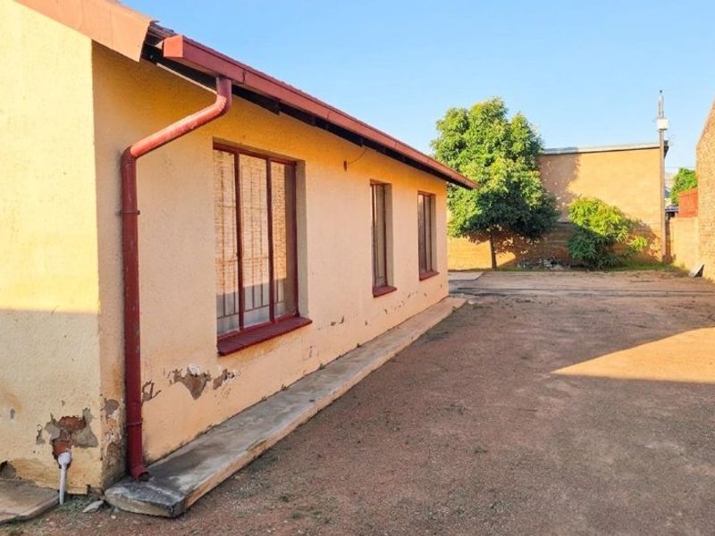 Rear exterior view of the property showing a plain facade with visible plaster damage and peeling paint, alongside a large open yard area.