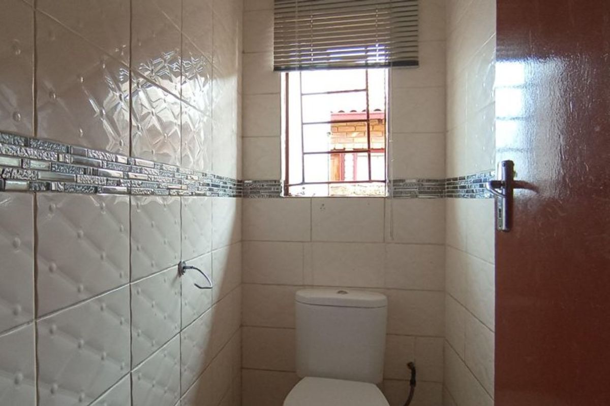A compact bathroom featuring full-height wall tiling, a standard toilet, and a window with security bars and a blind.