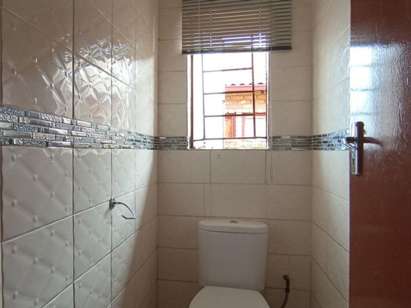 A compact bathroom featuring full-height wall tiling, a standard toilet, and a window with security bars and a blind.