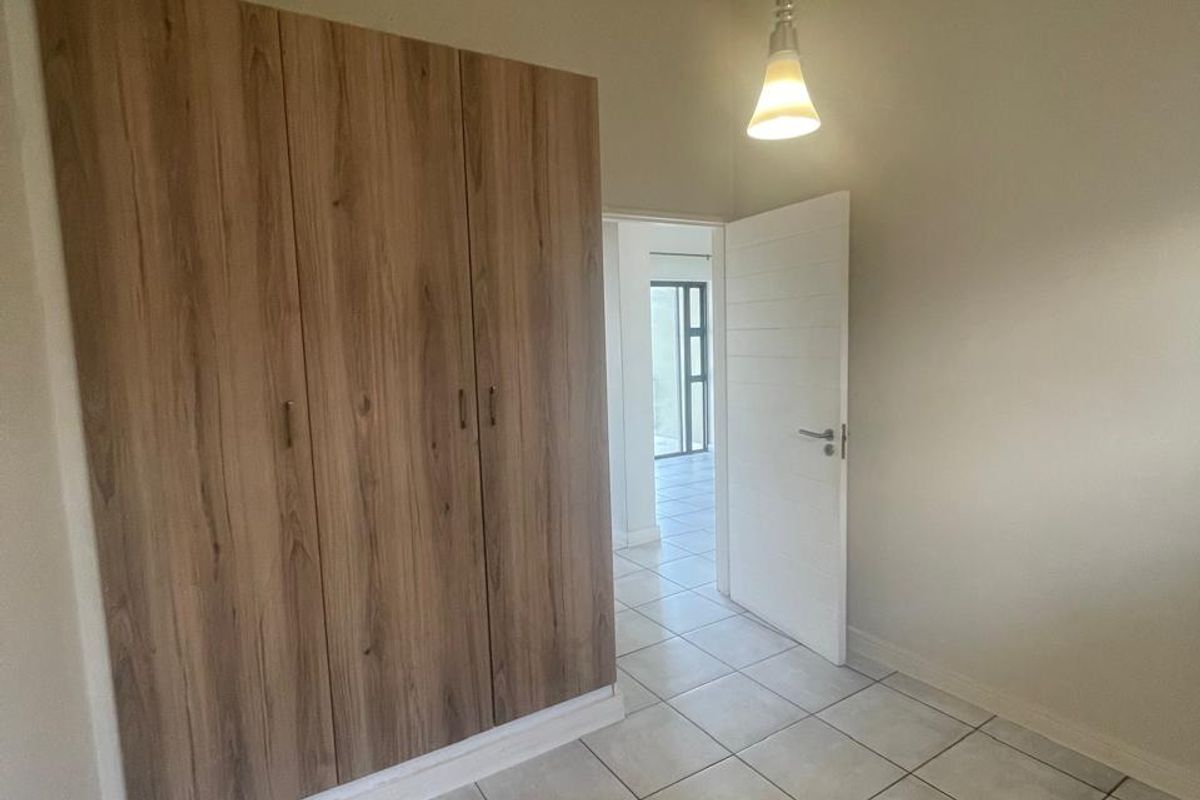 A clean, empty bedroom featuring modern wood-finish built-in cupboards, neutral tiled flooring, and a simple pendant light fixture.