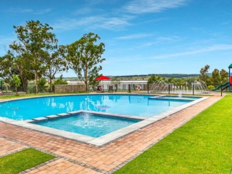 A well-maintained outdoor swimming pool area featuring a water fountain, paved surround, and landscaped lawn with perimeter fencing.