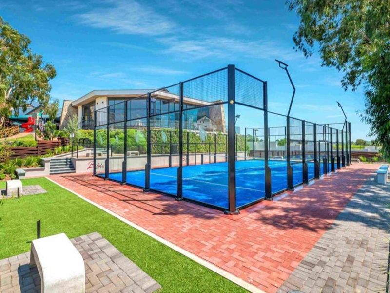 A well-maintained outdoor padel court facility featuring blue court surfacing, perimeter fencing, and landscaped paved surrounds.