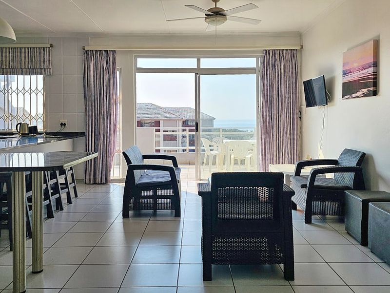 Open-plan living and kitchen area featuring tiled floors, a ceiling fan, and a sliding door leading to a balcony with coastal views.