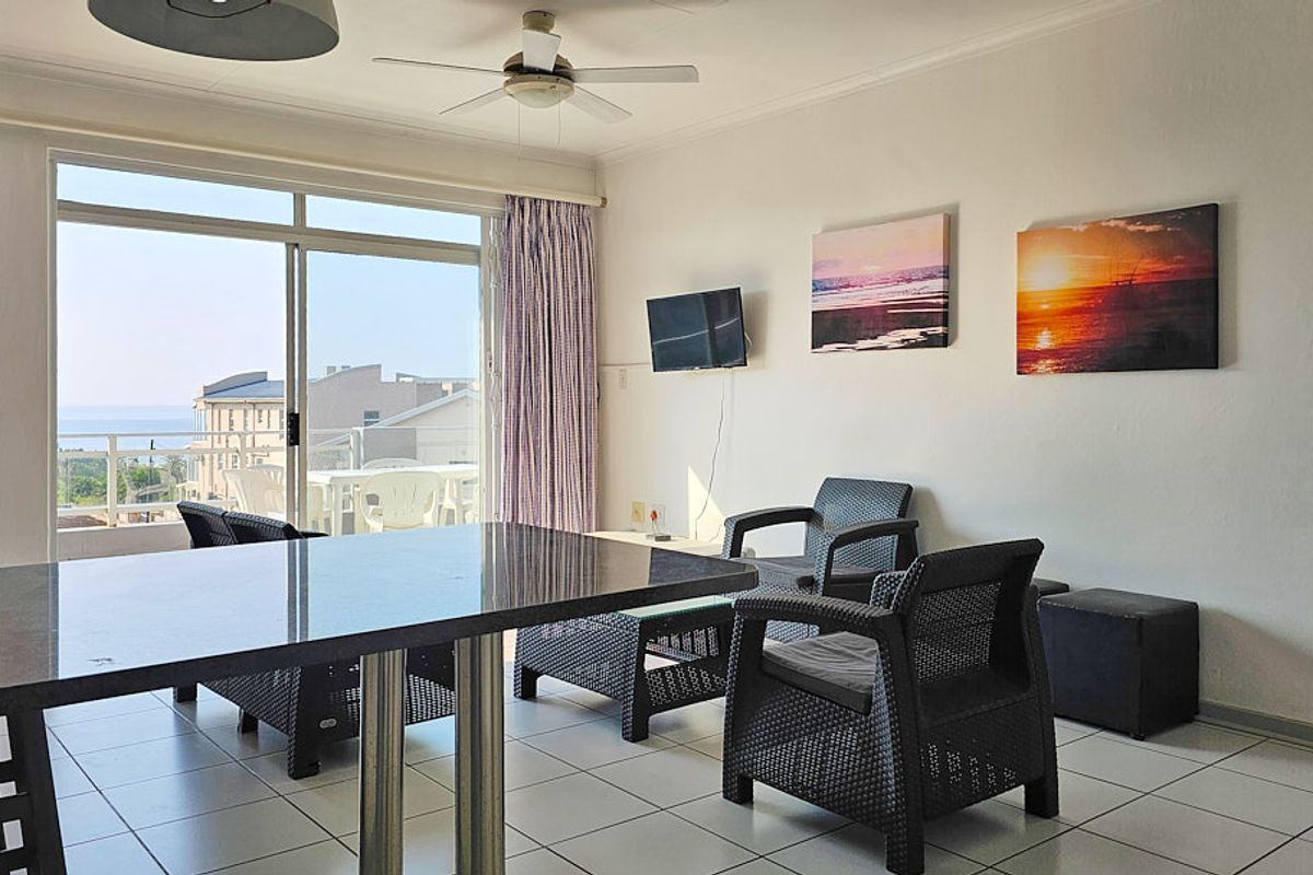 A bright living area featuring tiled flooring, a ceiling fan, and large sliding glass doors that open onto a balcony with coastal views.