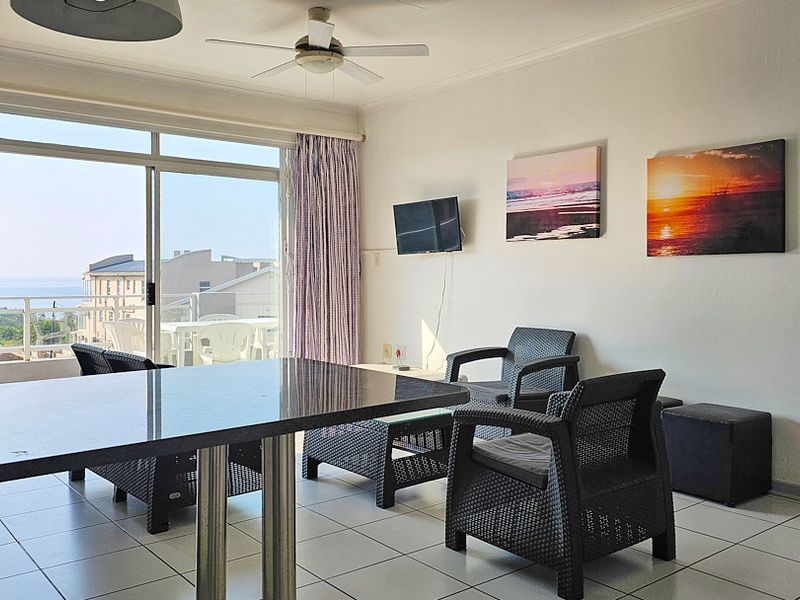 A bright living area featuring tiled flooring, a ceiling fan, and large sliding glass doors that open onto a balcony with coastal views.