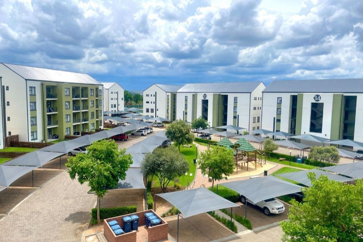 Aerial view of an apartment complex featuring multiple residential blocks, paved driveways, shade cloth parking bays, and a central landscaped garden with a play area.