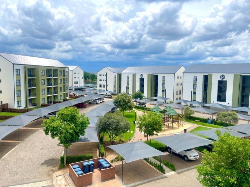 Aerial view of an apartment complex featuring multiple residential blocks, paved driveways, shade cloth parking bays, and a central landscaped garden with a play area.