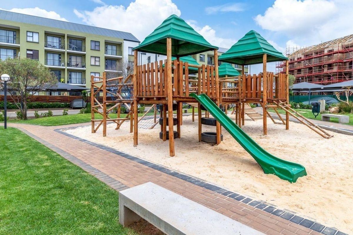 Communal playground area featuring wooden climbing structures, a slide, and a sand pit, set within a well-maintained apartment complex garden.