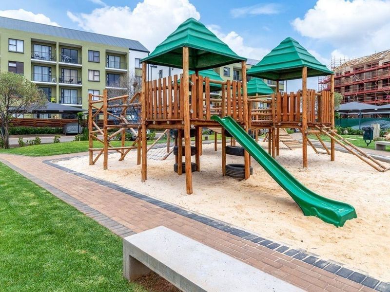 Communal playground area featuring wooden climbing structures, a slide, and a sand pit, set within a well-maintained apartment complex garden.