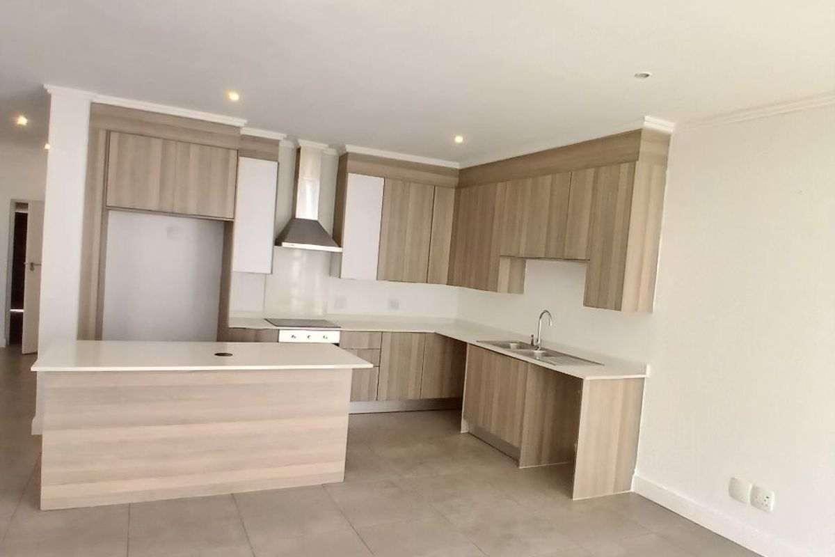 Modern open-plan kitchen featuring light wood-look cabinetry, stone countertops, stainless steel extractor fan, and neutral tiled flooring in a clean, vacant space.