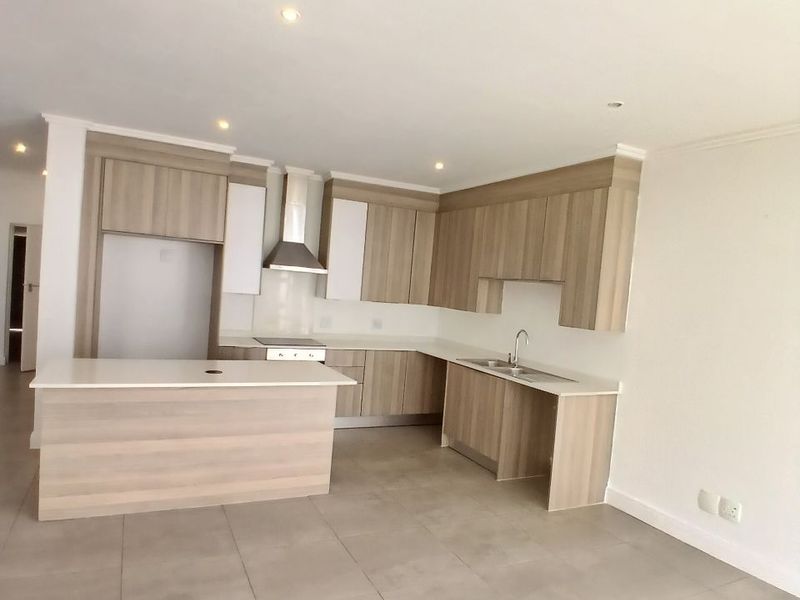 Modern open-plan kitchen featuring light wood-look cabinetry, stone countertops, stainless steel extractor fan, and neutral tiled flooring in a clean, vacant space.