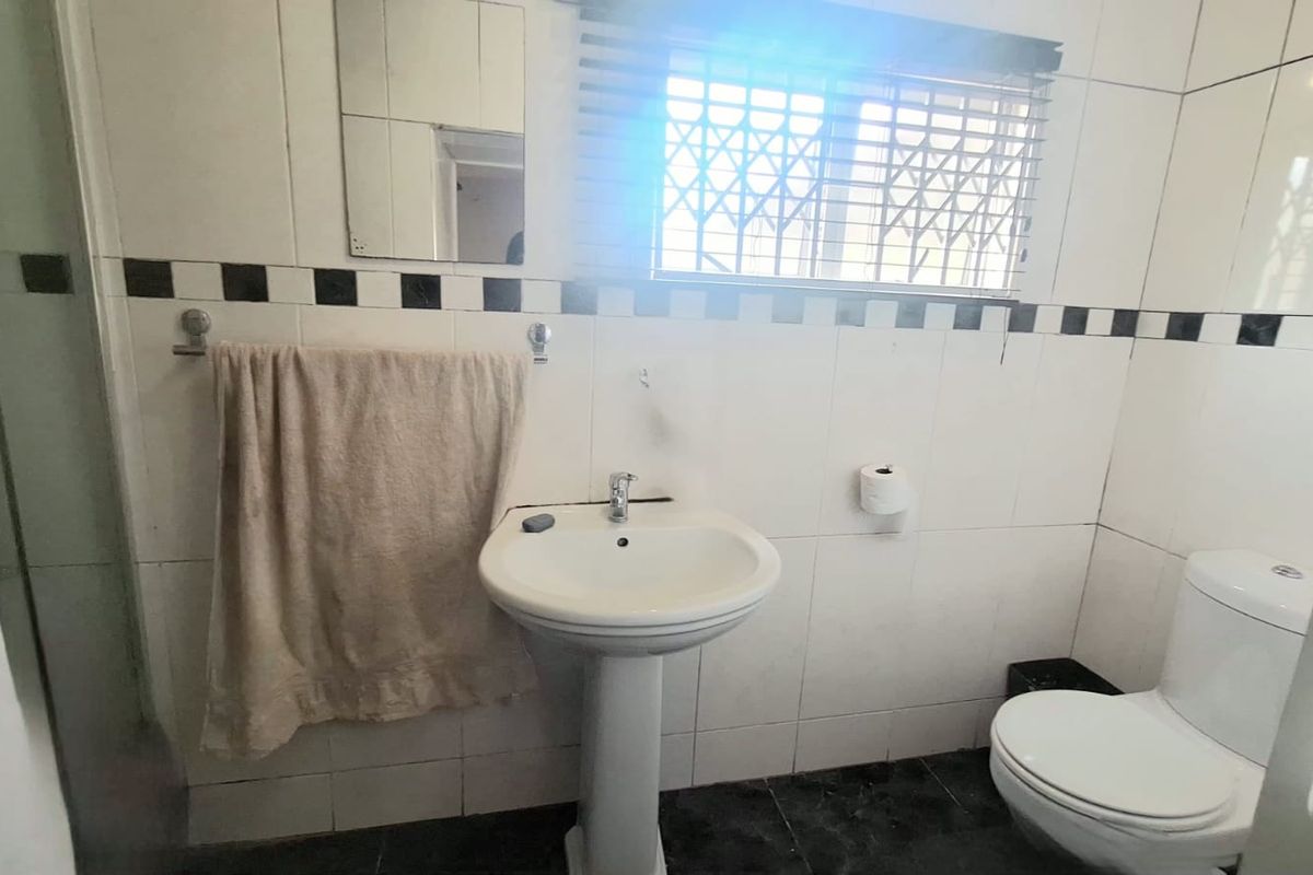 A functional bathroom featuring a pedestal basin, toilet, and shower enclosure with tiled finishes and a window with security bars.