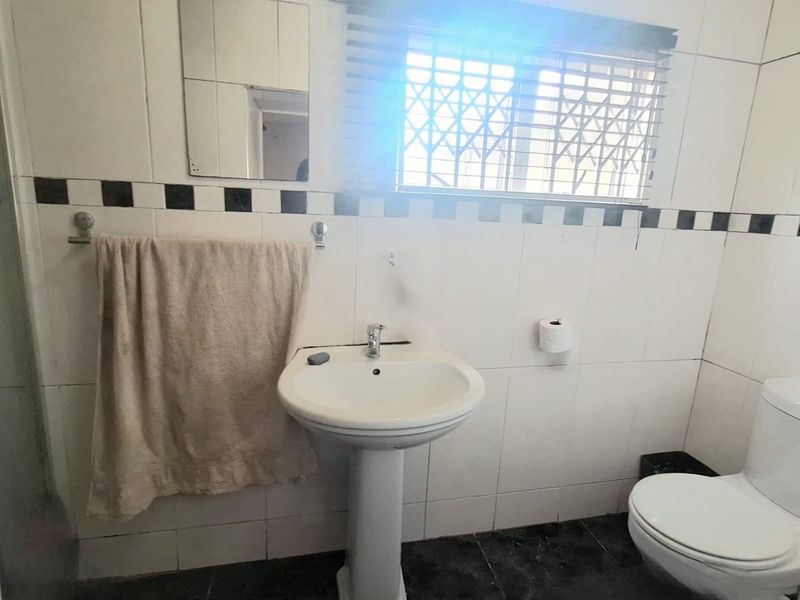 A functional bathroom featuring a pedestal basin, toilet, and shower enclosure with tiled finishes and a window with security bars.