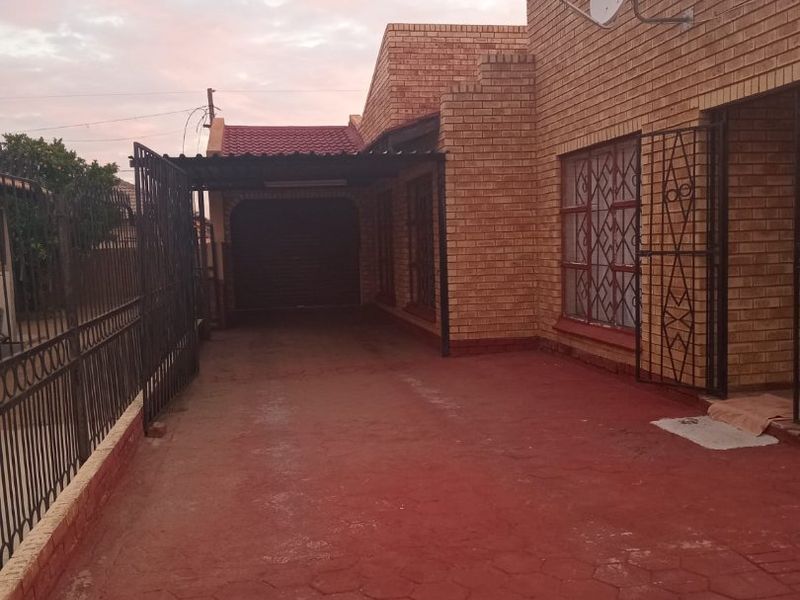 Front exterior view of a face brick house featuring a paved driveway, carport, and secure metal perimeter fencing.