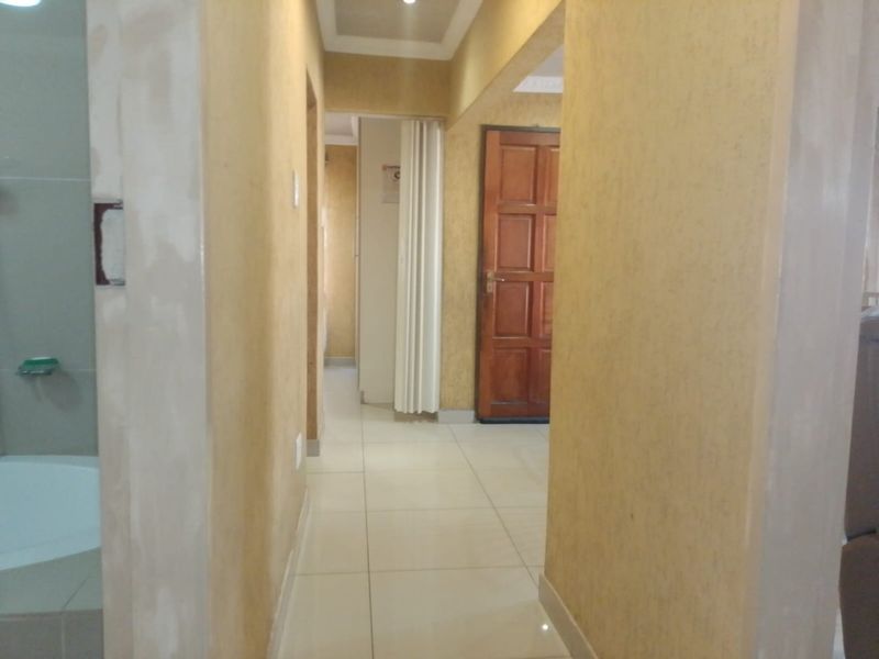 A narrow hallway featuring tiled flooring, recessed lighting, and a wooden door, currently showing signs of unfinished wall surfaces and general wear.