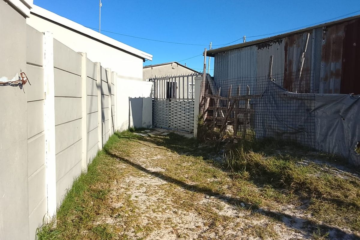 A rear exterior view showing a precast concrete boundary wall, a metal security gate, and a corrugated metal structure with visible signs of wear.