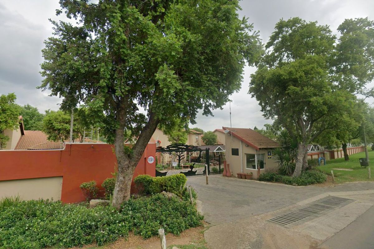 Front entrance view of the apartment complex featuring a security gate, guard house, and perimeter wall with mature trees.
