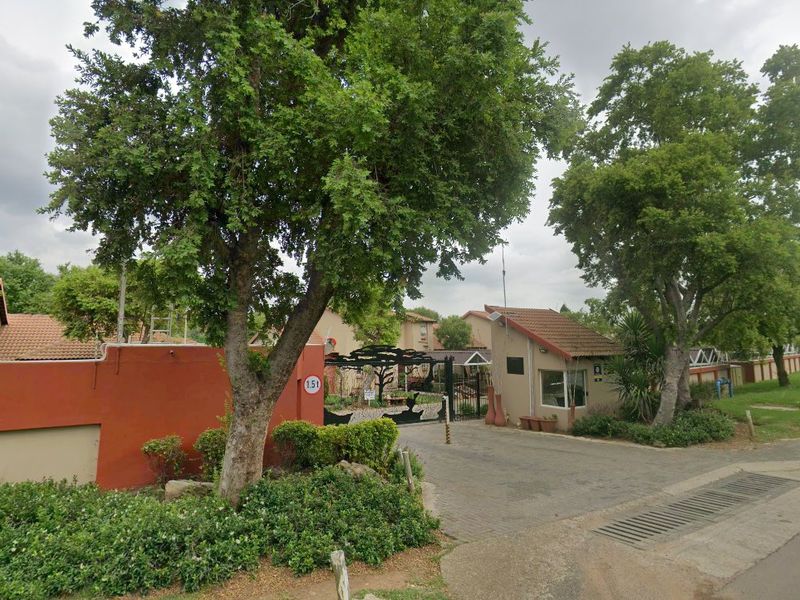 Front entrance view of the apartment complex featuring a security gate, guard house, and perimeter wall with mature trees.