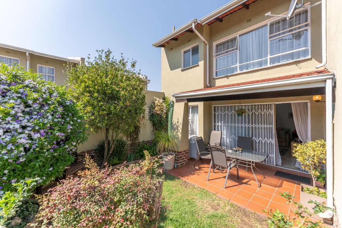 A rear exterior view of the townhouse featuring a tiled patio area, established garden, and sliding door access with security gate.