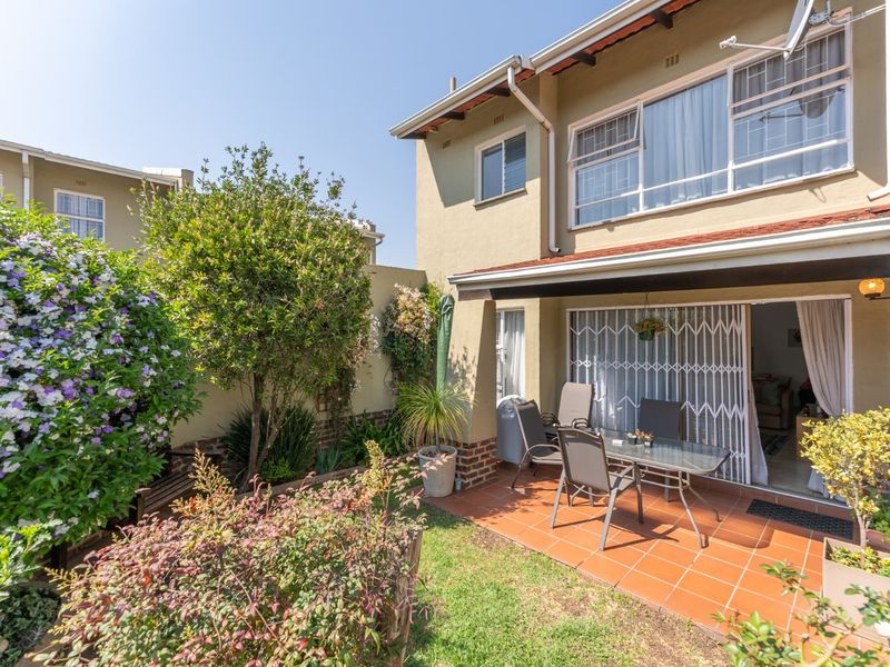 A rear exterior view of the townhouse featuring a tiled patio area, established garden, and sliding door access with security gate.