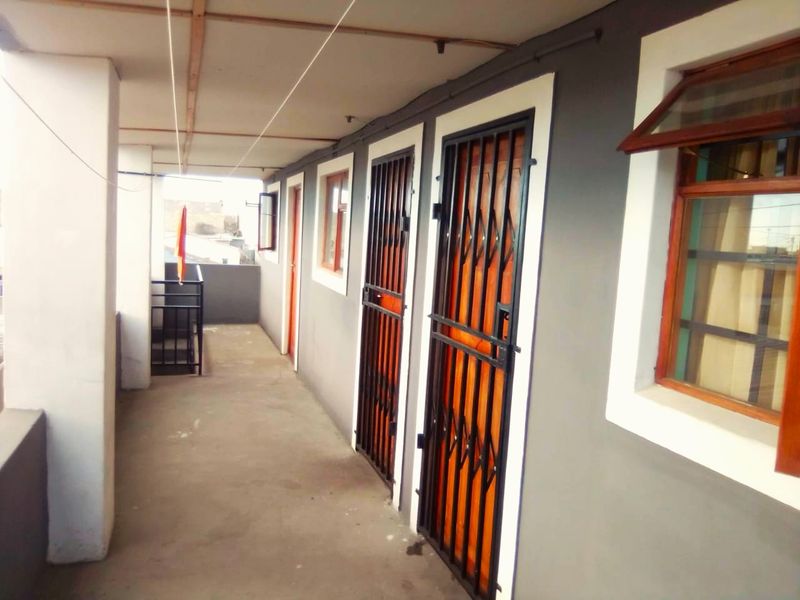 A covered exterior walkway or balcony featuring concrete flooring, security gates, and timber frame windows along the building facade.
