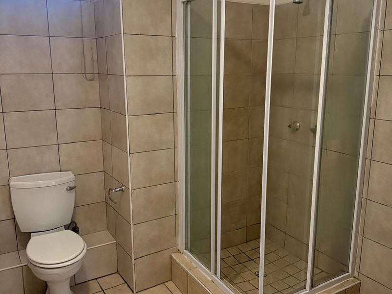 A functional bathroom featuring a corner shower enclosure, a standard toilet, and neutral floor-to-ceiling wall tiling.