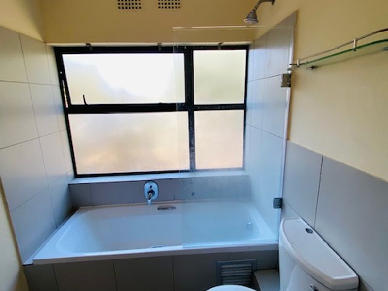 A functional bathroom featuring a built-in bathtub with shower attachment, glass screen, toilet, and neutral wall tiling.