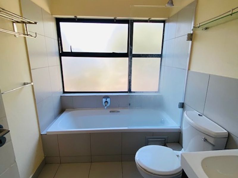 A functional bathroom featuring a built-in bathtub with shower head, toilet, and vanity basin, finished with neutral floor and wall tiling.