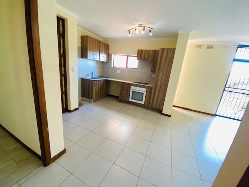 An open-plan kitchen and living area featuring modern wood-finish cabinetry, built-in oven and hob, tiled flooring, and a secure entrance door.
