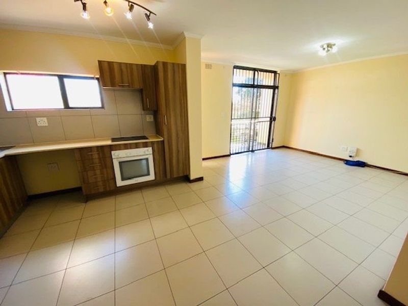 Spacious open-plan kitchen and living area featuring tiled flooring, modern wood-finish cabinetry, built-in oven and hob, and access to a balcony via a security gate.