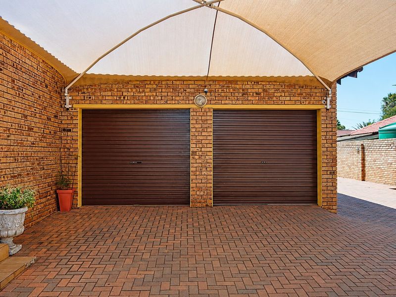 A well-maintained double garage featuring brown roller doors, brick exterior walls, and a paved driveway protected by a large shade sail.