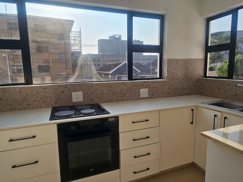 A bright, modern kitchen featuring stone countertops, a built-in oven with electric hob, and a double sink, complemented by large windows with city views.