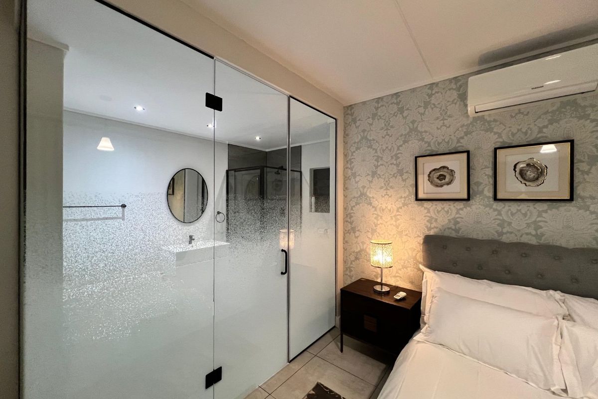 A modern bedroom featuring a glass-partitioned en-suite bathroom, tiled flooring, patterned wallpaper, and a wall-mounted split-system air conditioner.