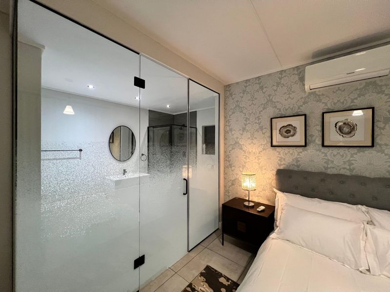 A modern bedroom featuring a glass-partitioned en-suite bathroom, tiled flooring, patterned wallpaper, and a wall-mounted split-system air conditioner.