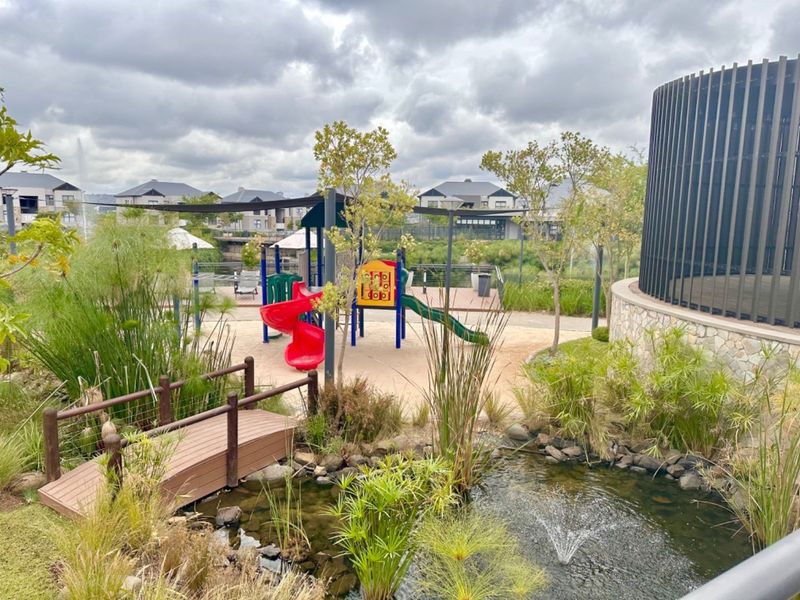 A well-maintained communal playground area featuring a water feature, wooden footbridge, and landscaped gardens within the apartment complex.