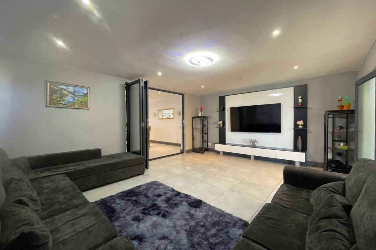 Spacious living room featuring modern tiled flooring, recessed downlighting, and a sleek built-in media wall unit with bi-fold doors leading to an adjacent room.