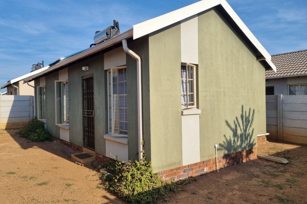 Rear view of a single-story house featuring a solar water heater, security gate, and brick plinth foundation with surrounding open yard space.