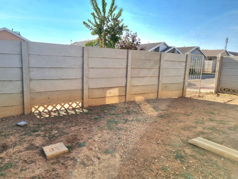 A rear yard area featuring a precast concrete boundary wall, a metal pedestrian gate, and an unlandscaped dirt ground surface.