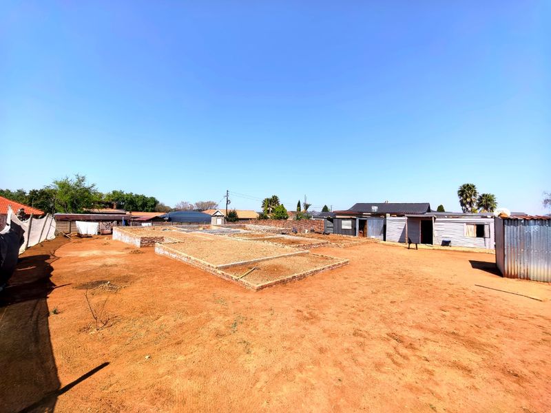 A vacant residential plot featuring an unfinished brick foundation and existing corrugated metal structures on a level, unpaved site.