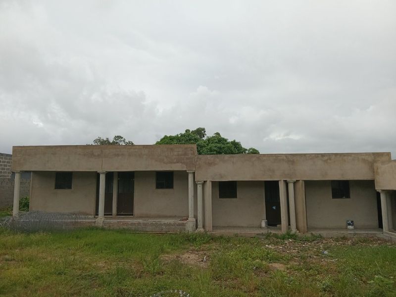 A single-story residential building exterior featuring an unfinished cement facade, covered veranda with concrete columns, and metal-framed windows set against a grassy yard.