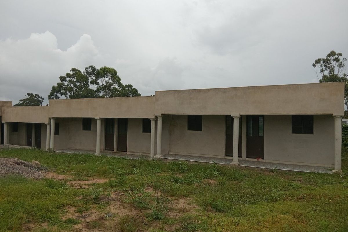 Exterior view of a single-story apartment block featuring a covered veranda with concrete columns and unfinished exterior wall surfaces.